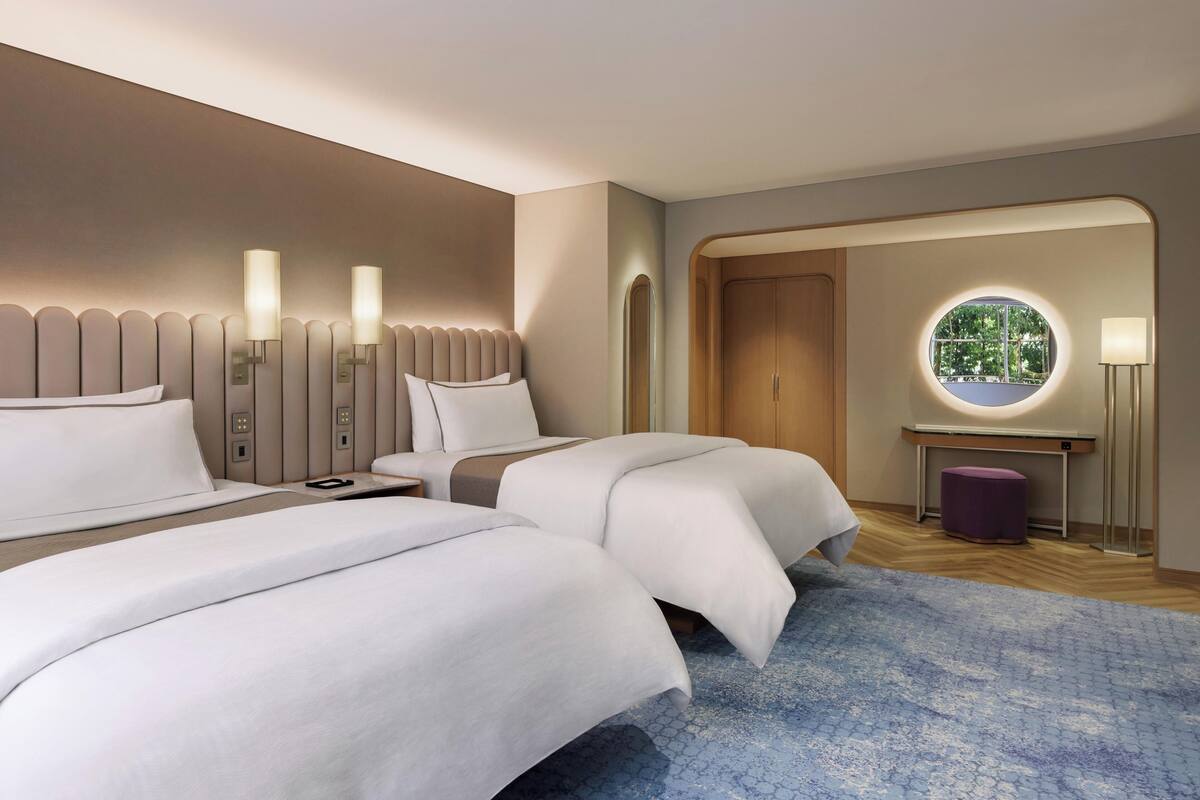 A hotel room with two beds, a round window, a nightstand, and a lamp.