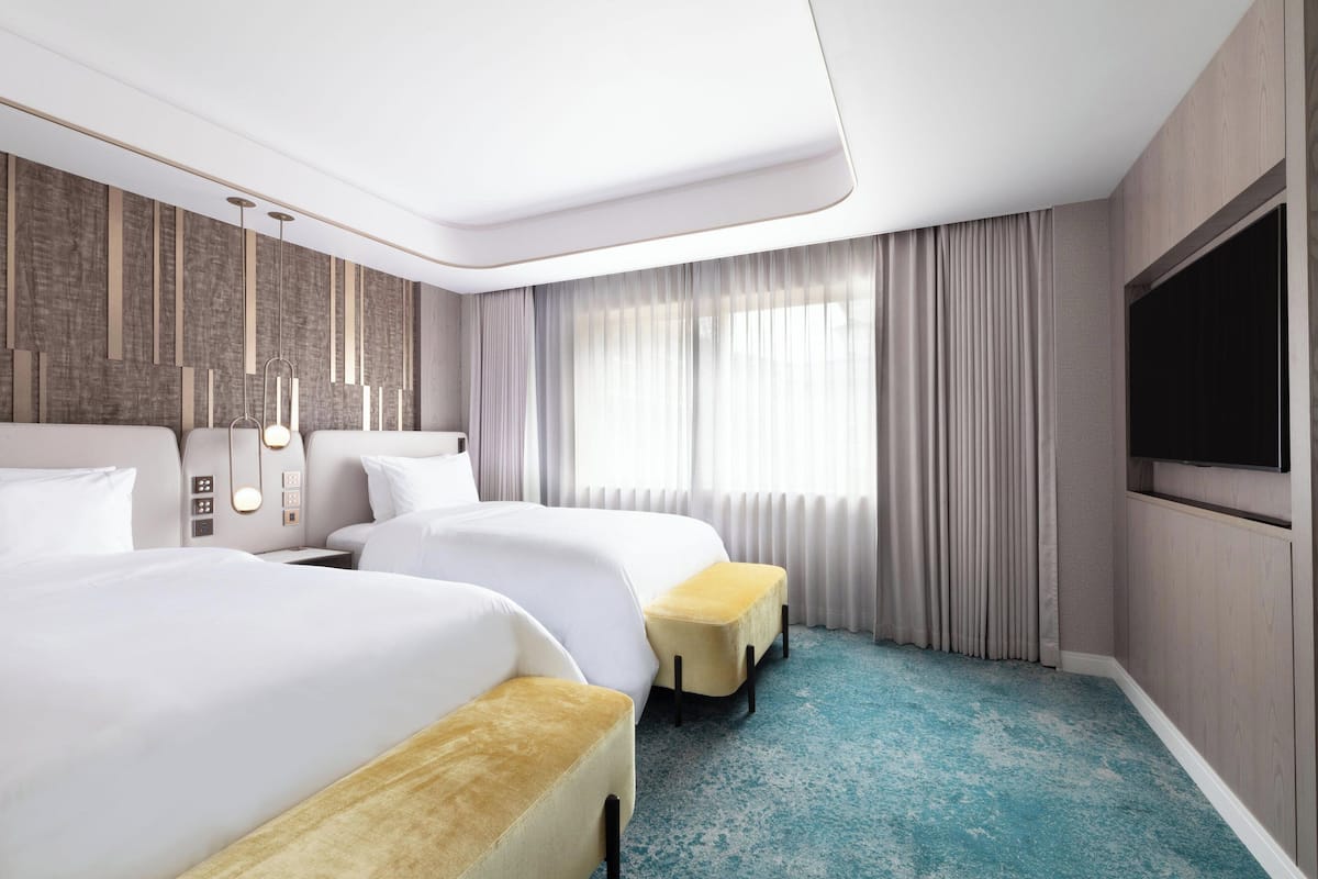 A modern hotel room with two beds, a flat-screen TV, a large window with curtains, and a unique blue carpet.