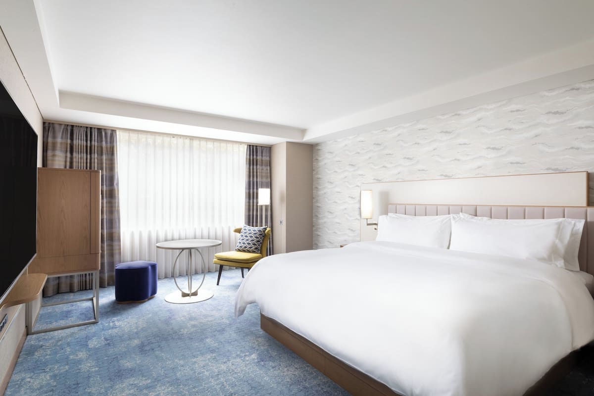 A hotel room with a large bed, a TV, a small round table, a chair, and a window with curtains.