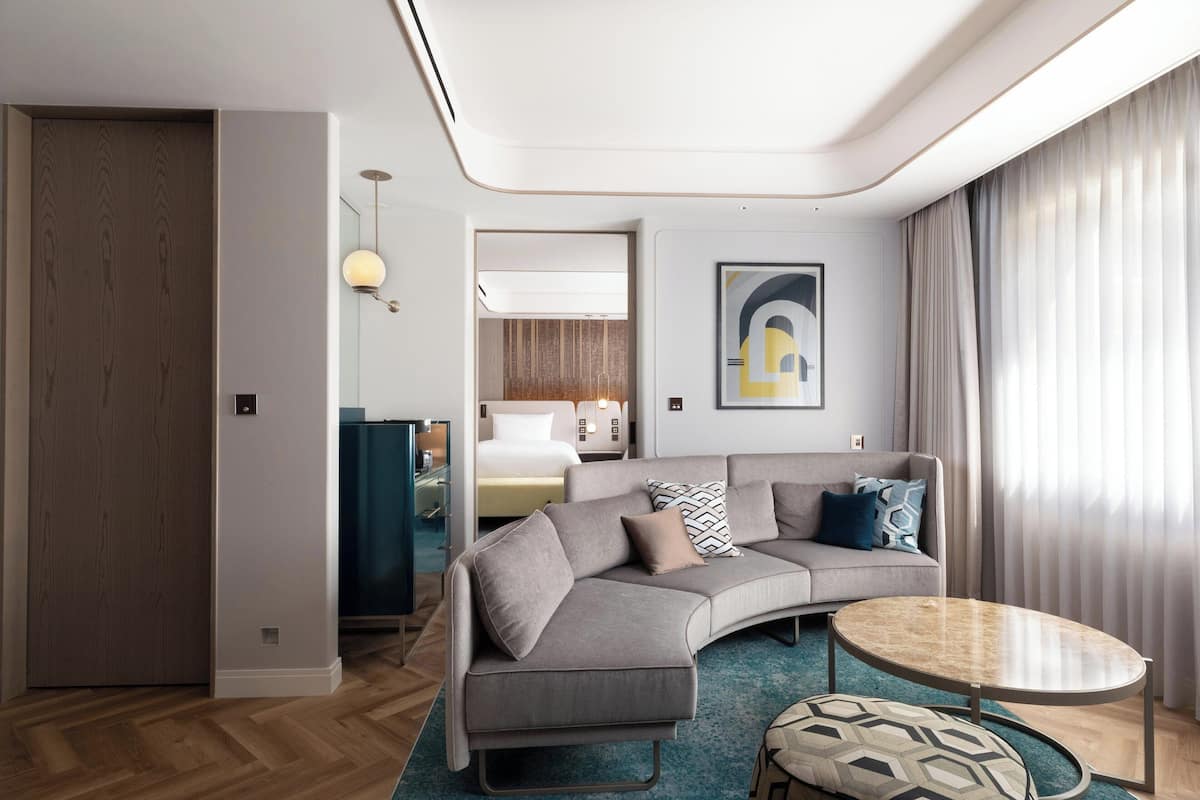 A modern hotel room with a curved gray sofa, a round coffee table, and a large window with curtains.