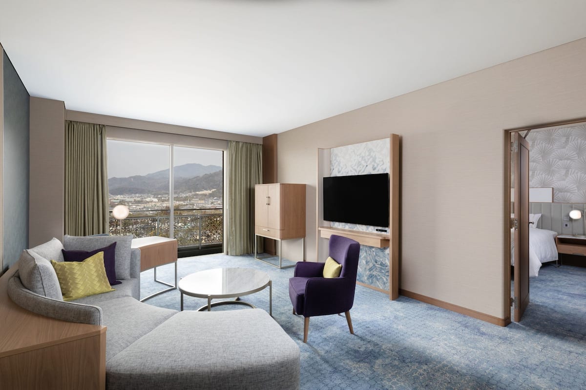 A modern hotel room with a large window, a flat-screen TV, a sofa, a chair, and a small table.
