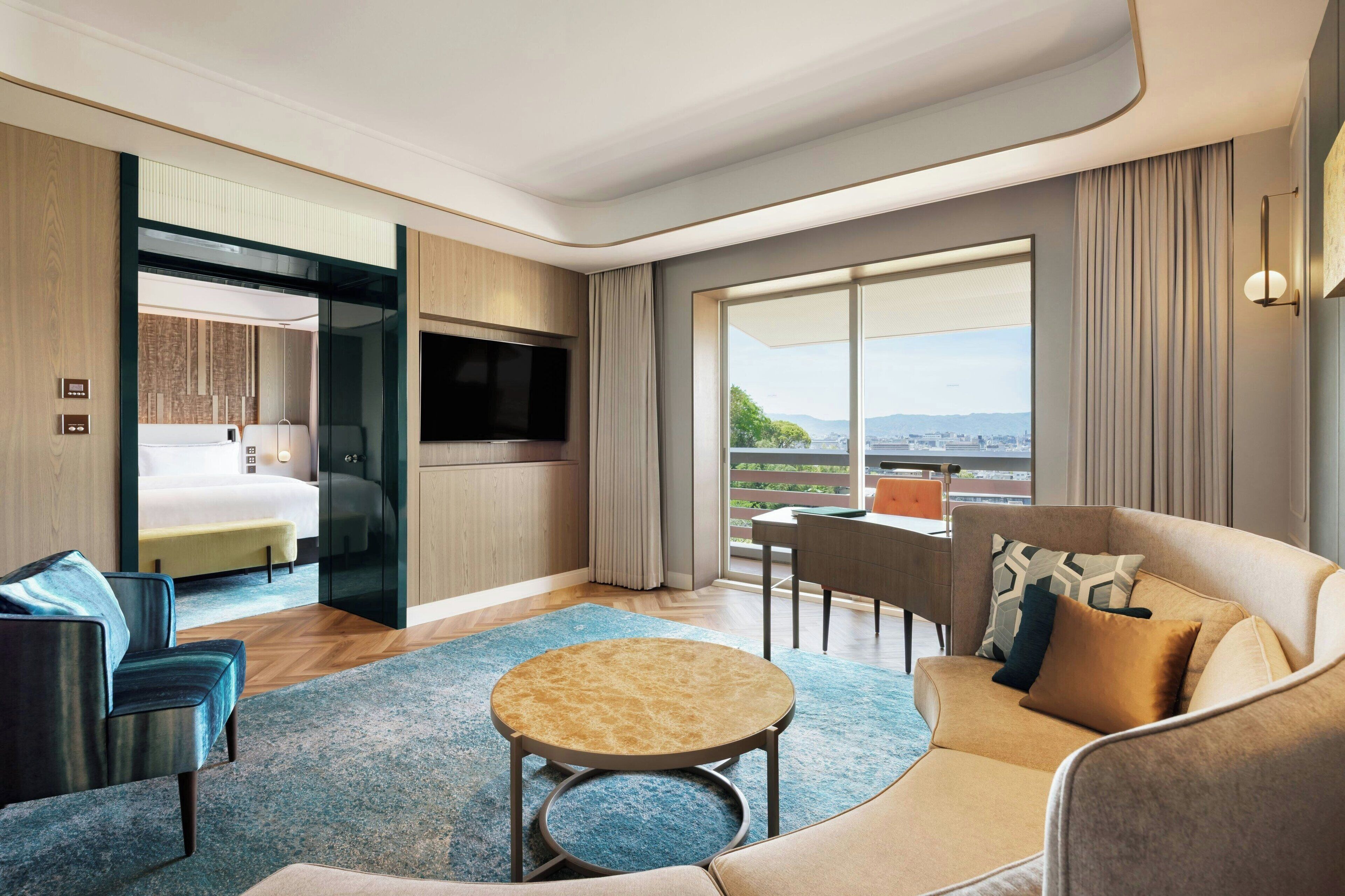 Una habitación de hotel moderna con sofá, una mesita redonda, televisor, cama y balcón con vistas.