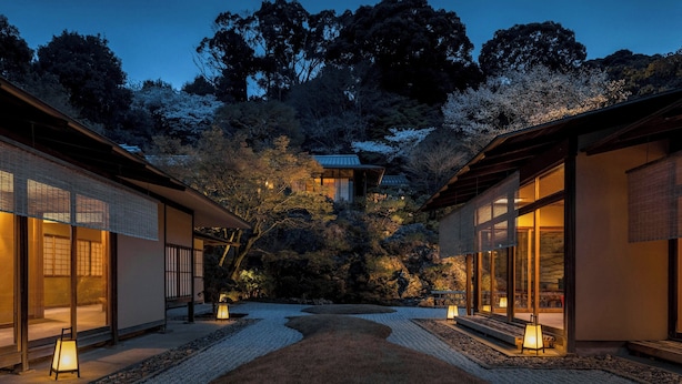 Un edificio tradicional japonés de madera con grandes ventanales, rodeado por un jardín con un sendero de grava y faroles.
