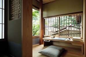 A traditional Japanese room with tatami flooring, a low wooden desk with a laptop, a small cushion, and a window with a view of a garden and a wooden bridge.