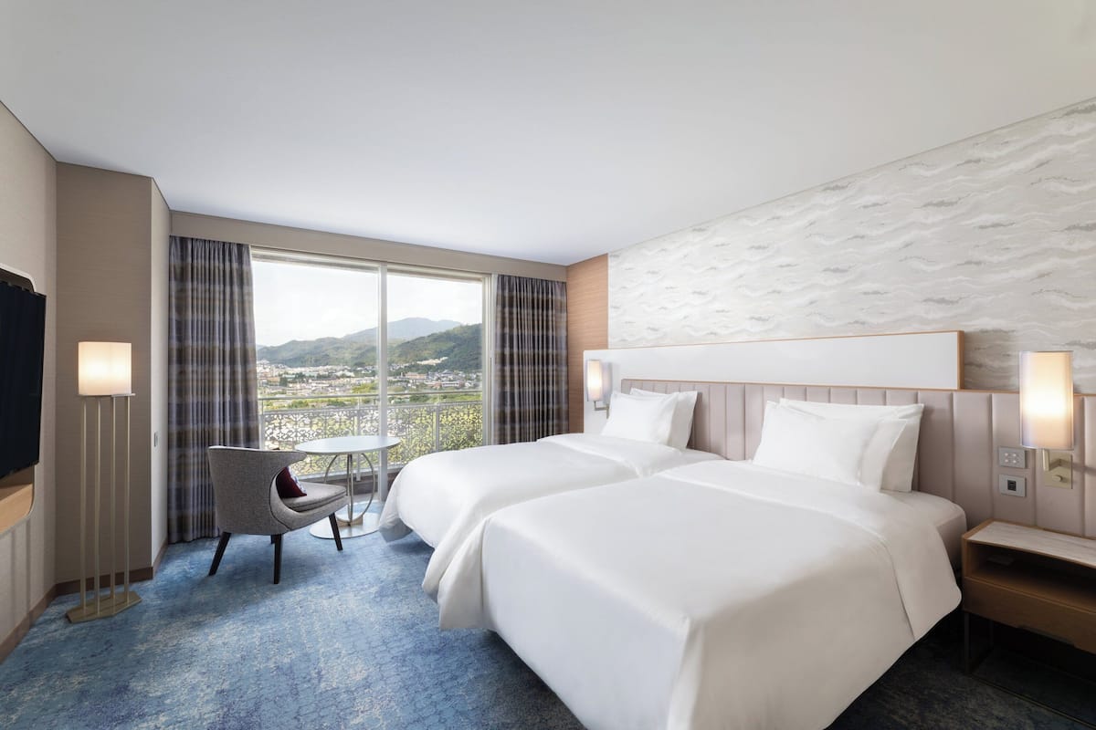 A hotel room with two beds, a small table, a chair, and a view of the city and mountains.