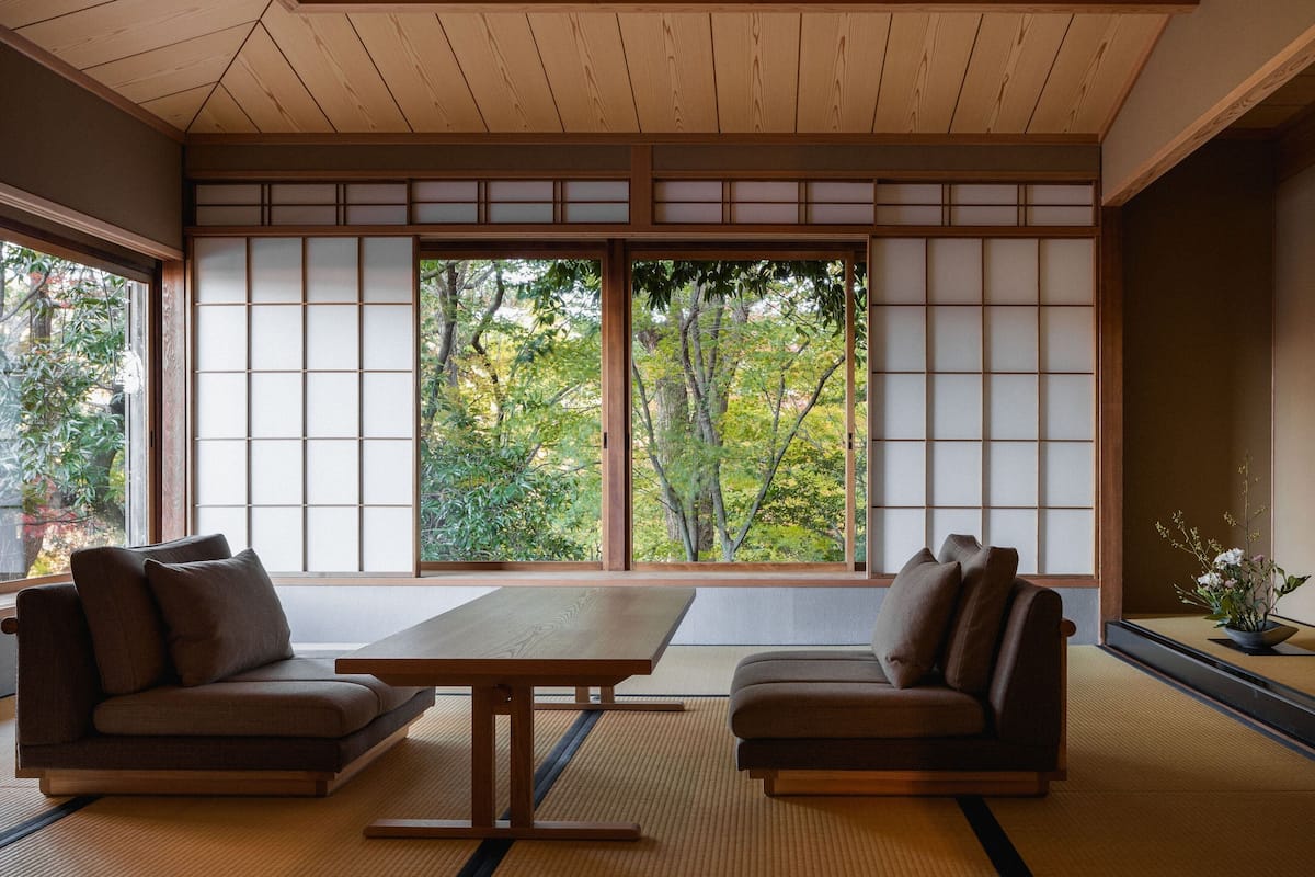 A traditional Japanese room with tatami flooring, a low table, two sofas, and sliding doors with a view of trees.