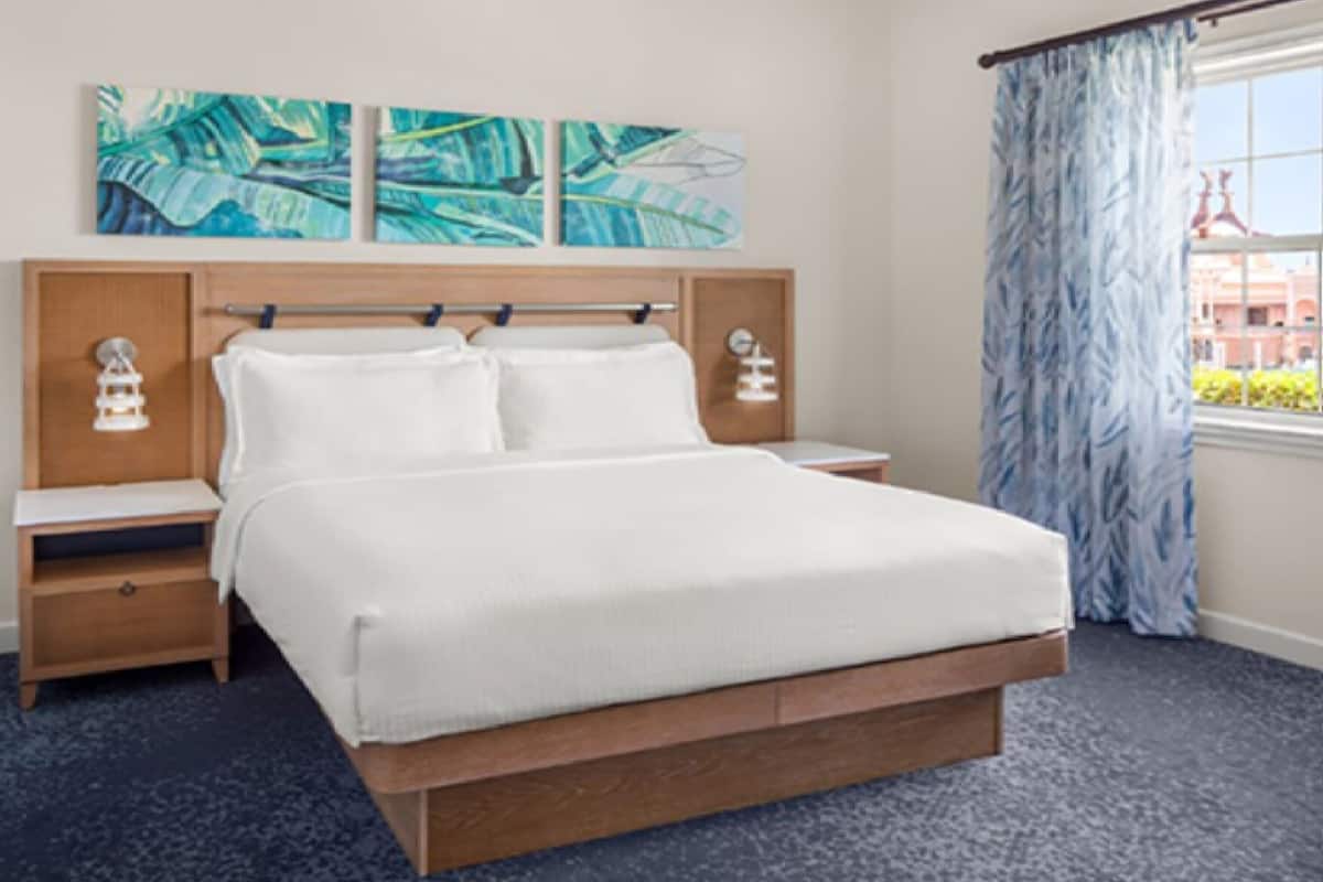 A hotel room with a large bed, wooden headboard, two bedside tables, and a view of a building through a window with blue curtains.