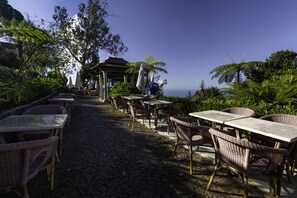 A cobblestone pathway leading to outdoor seating with wicker chairs and tables, surrounded by lush greenery and overlooking a scenic view. - Quinta do Monte Panoramic Gardens (Funchal)