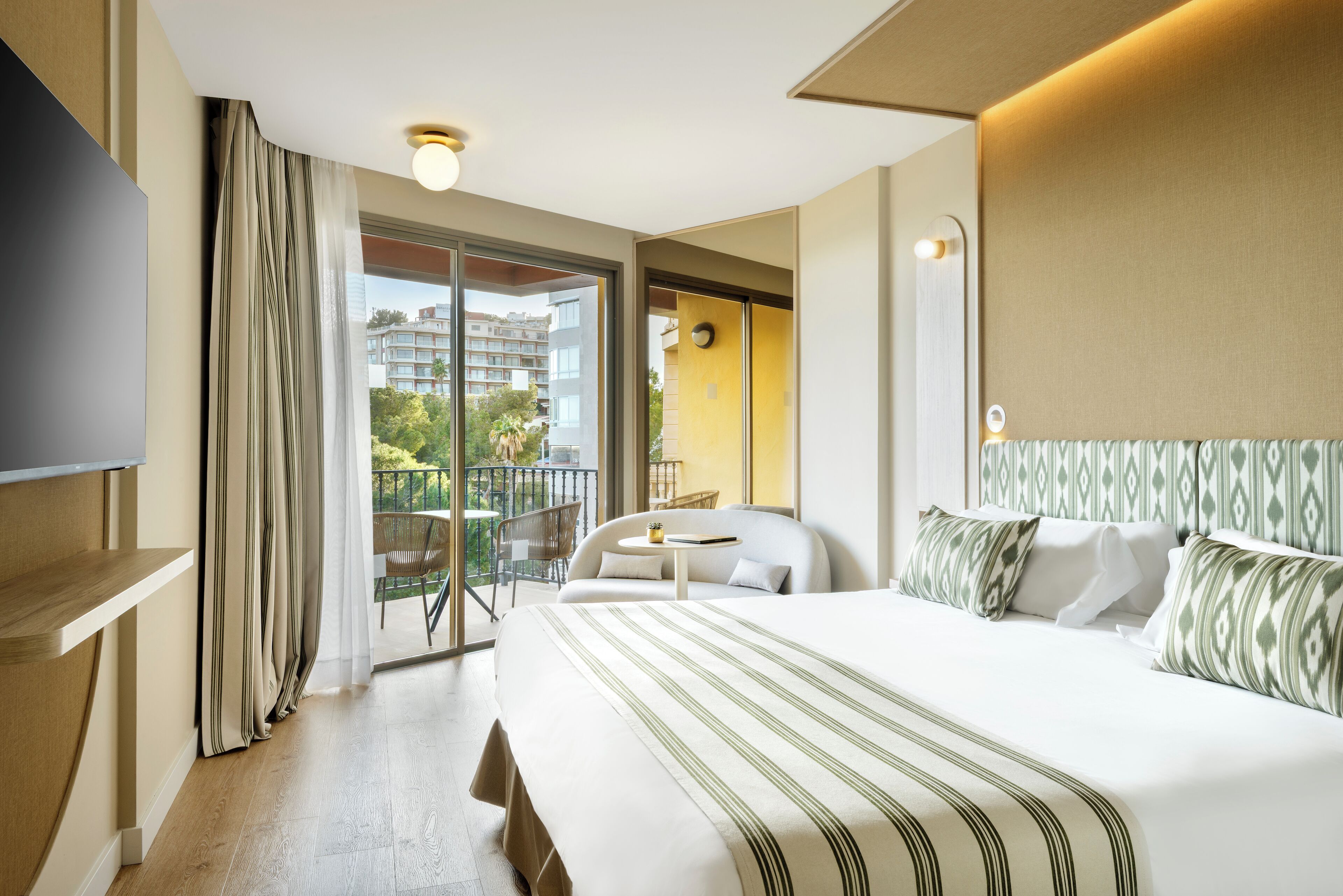 A hotel room with a large bed, a TV, a balcony with a chair, and a view of buildings.
