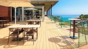 Un espacio exterior para comer con mesas y sillas, barandilla de vidrio y vista al océano.