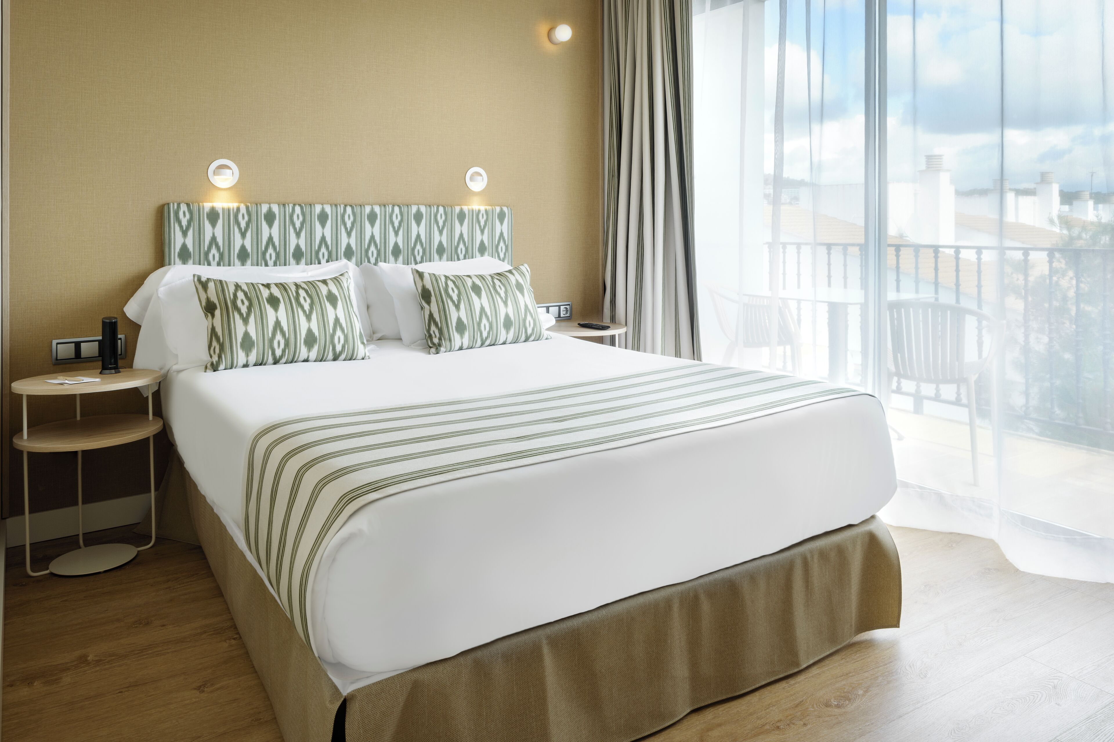 A hotel room with a large bed, two bedside tables, a nightstand, and a view of the city through large windows.