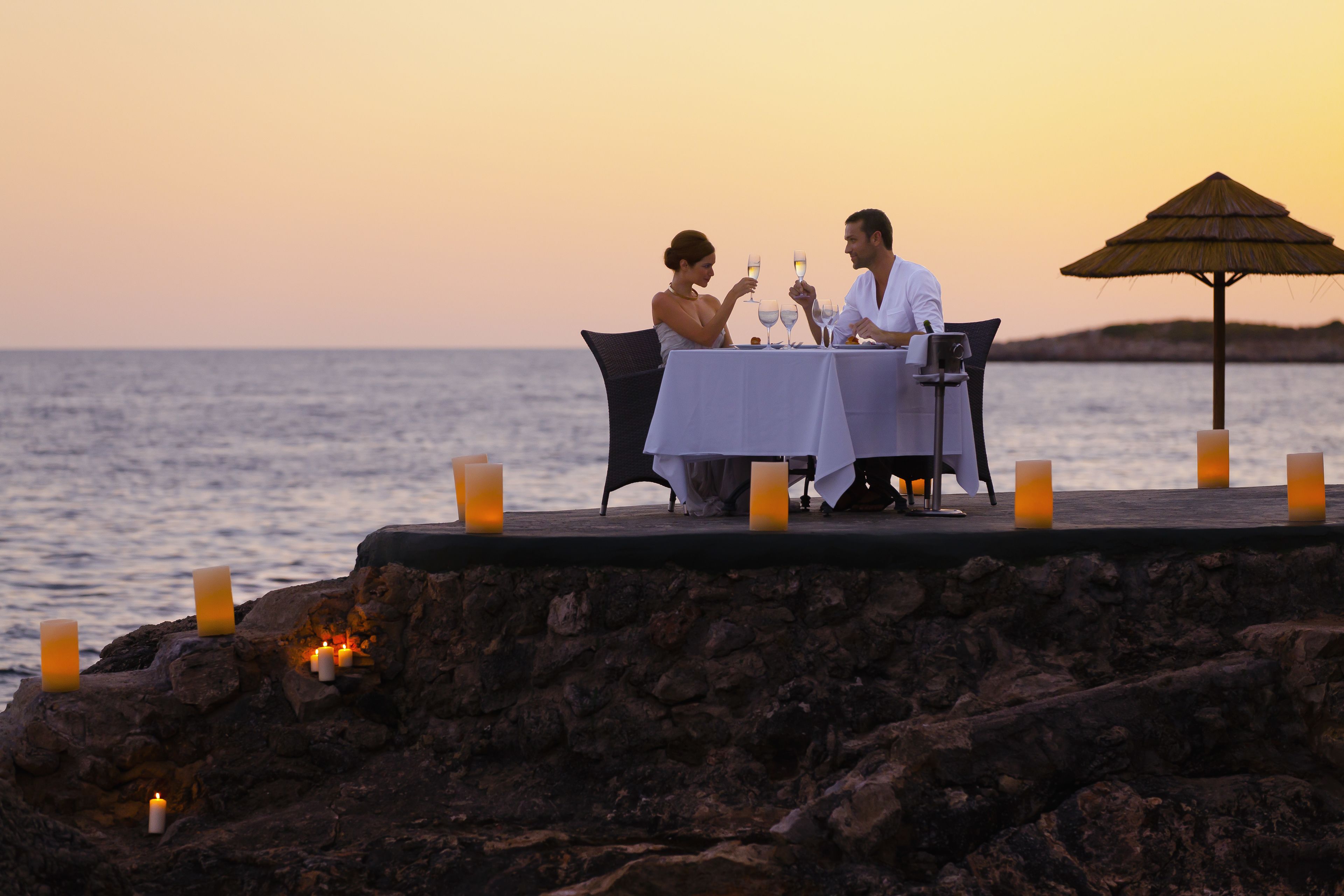 A couple dining at a table with candles on a rocky seaside terrace.
