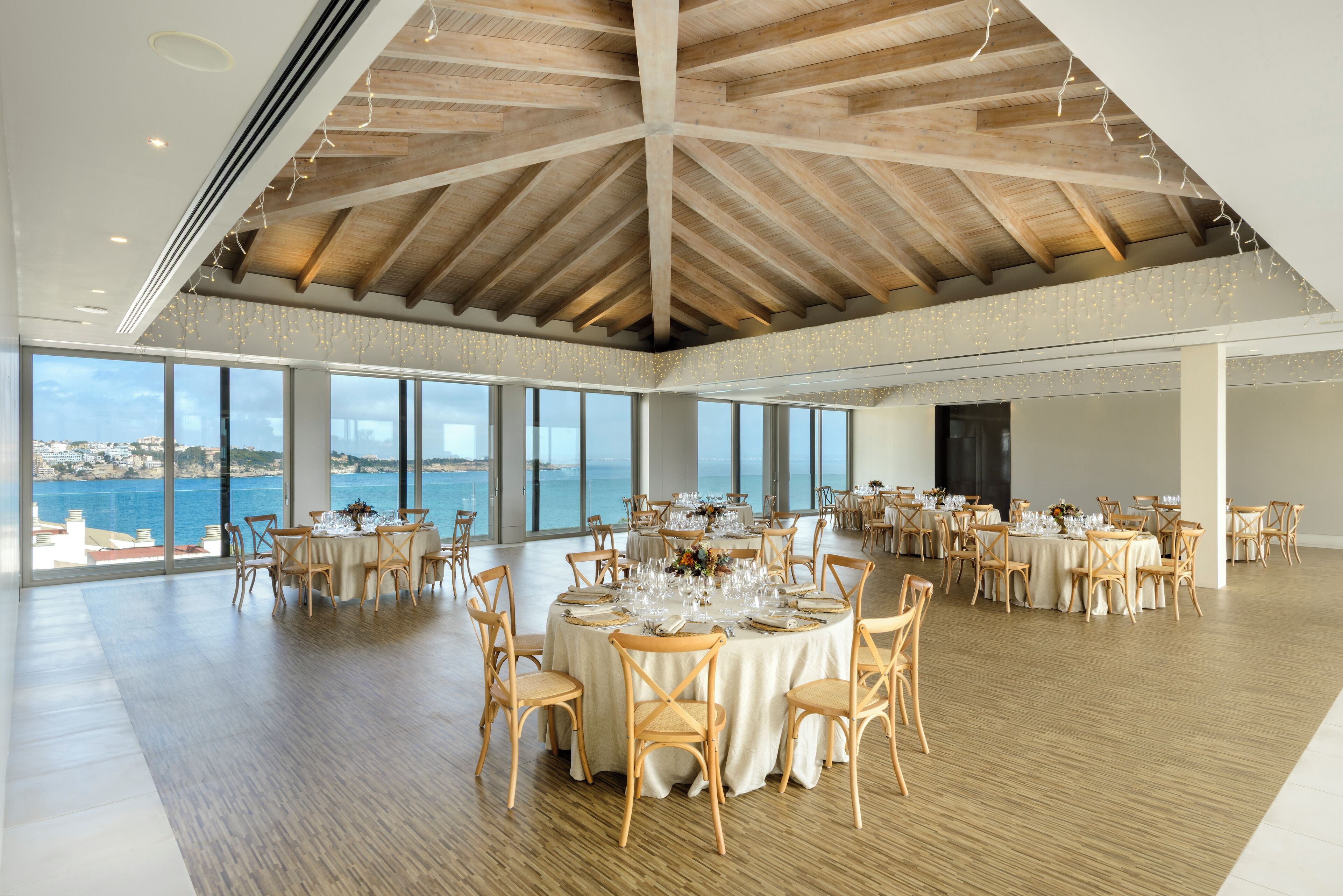 A spacious dining area with round tables set for a meal, large windows offering a view of the sea, and a high wooden ceiling.