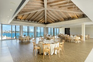 A spacious dining area with round tables set for a meal, large windows offering a view of the sea, and a high wooden ceiling. - Barceló Illetas Albatros - Adults Only (Calvia)