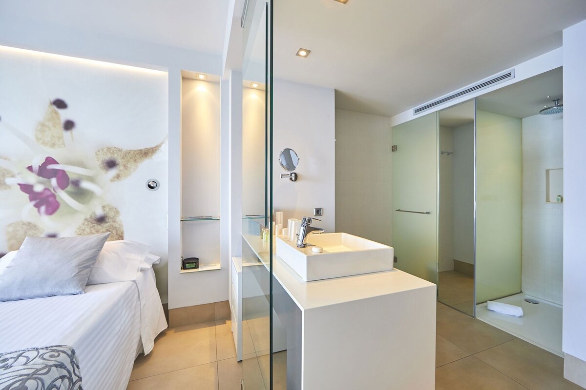 A modern bedroom with a bed, a white bedside table, a glass partition, a bathroom with a glass shower enclosure, and a wall-mounted mirror.