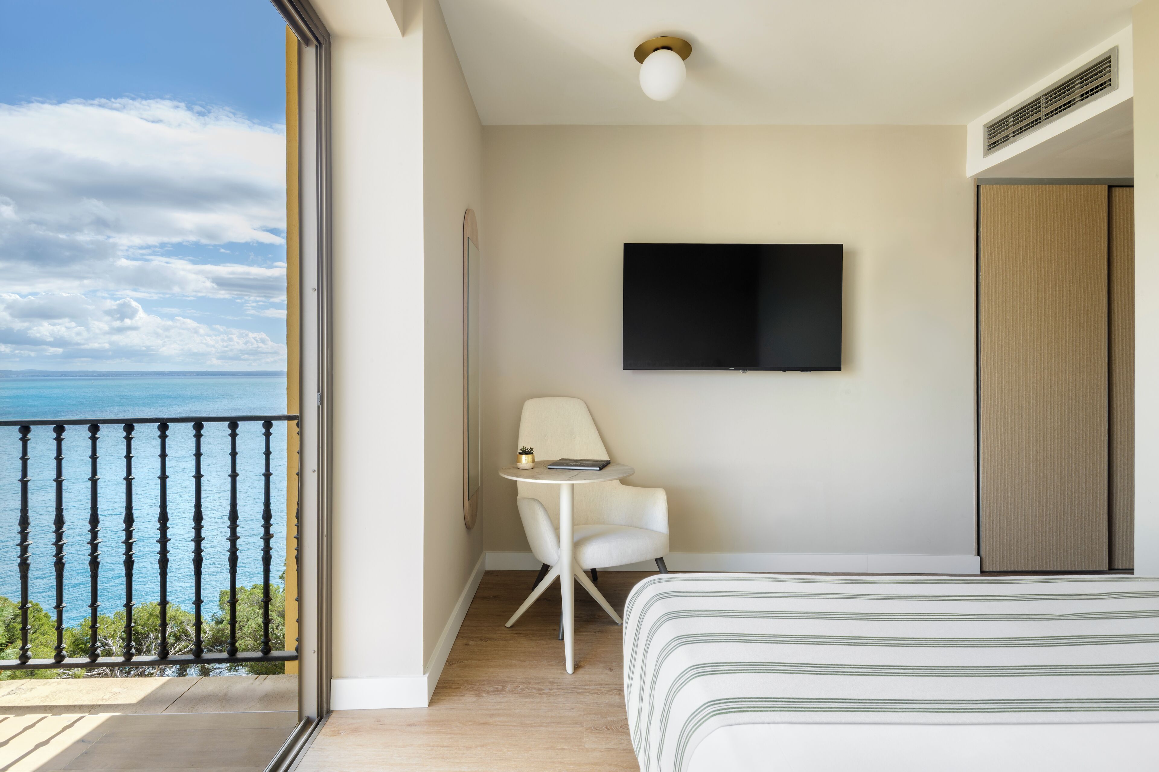 Habitación de hotel con cama, televisión montada en la pared, una mesita con silla y balcón con vista al mar.