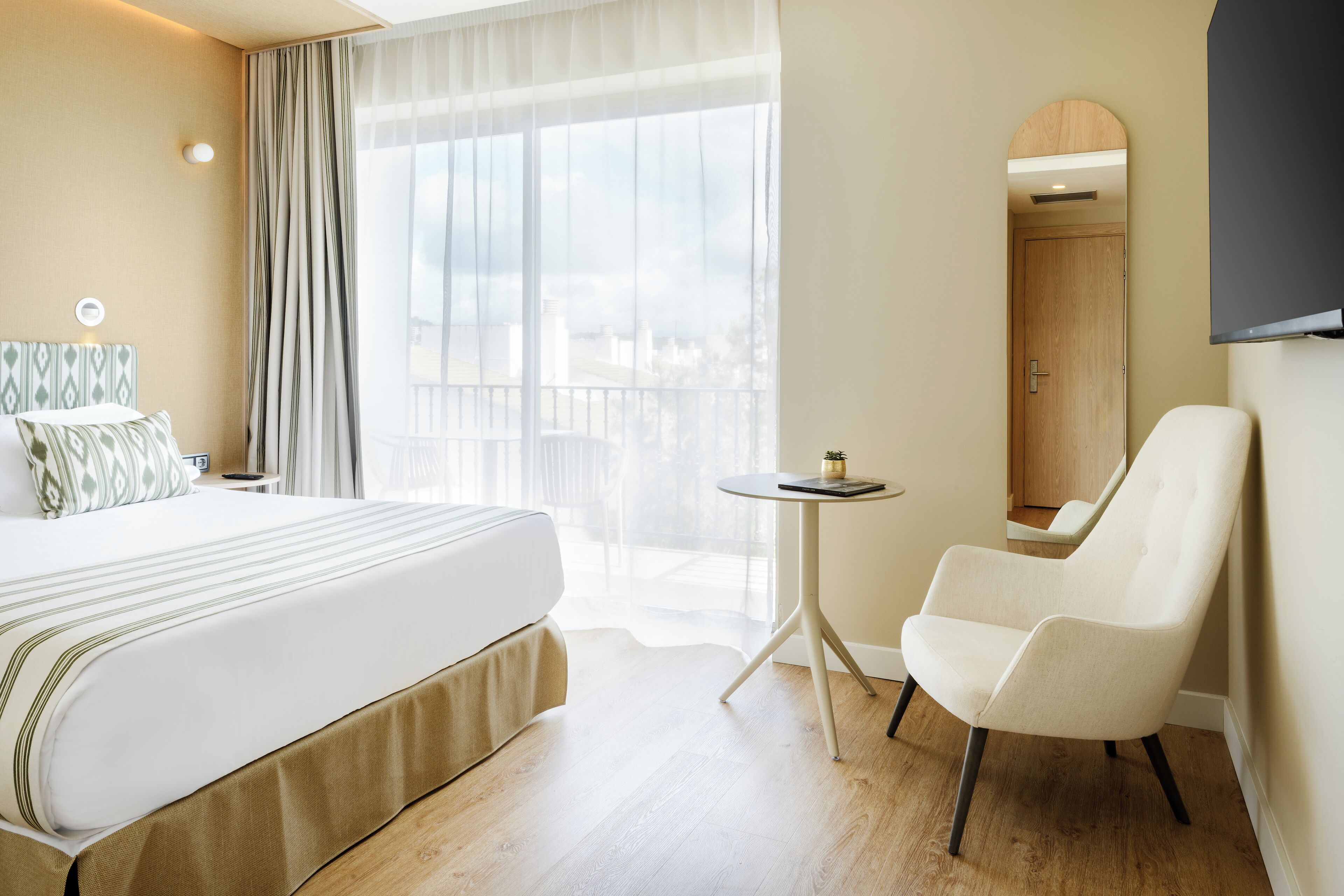 Une chambre d’hôtel avec un grand lit, une petite table ronde, une chaise, un miroir et une vue sur la ville par la fenêtre.