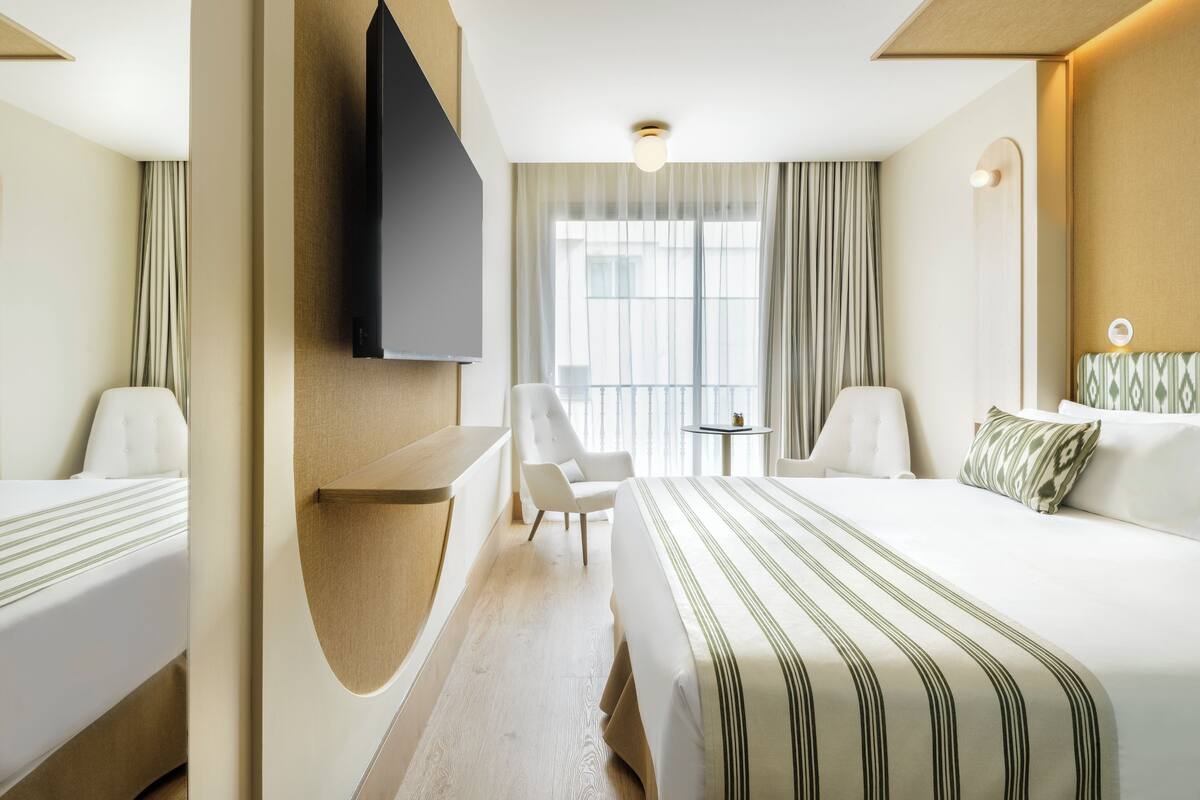 Hotellihuone, jossa on sänky, tuoli, työpöytä, televisio ja ikkuna, jossa on verhot.