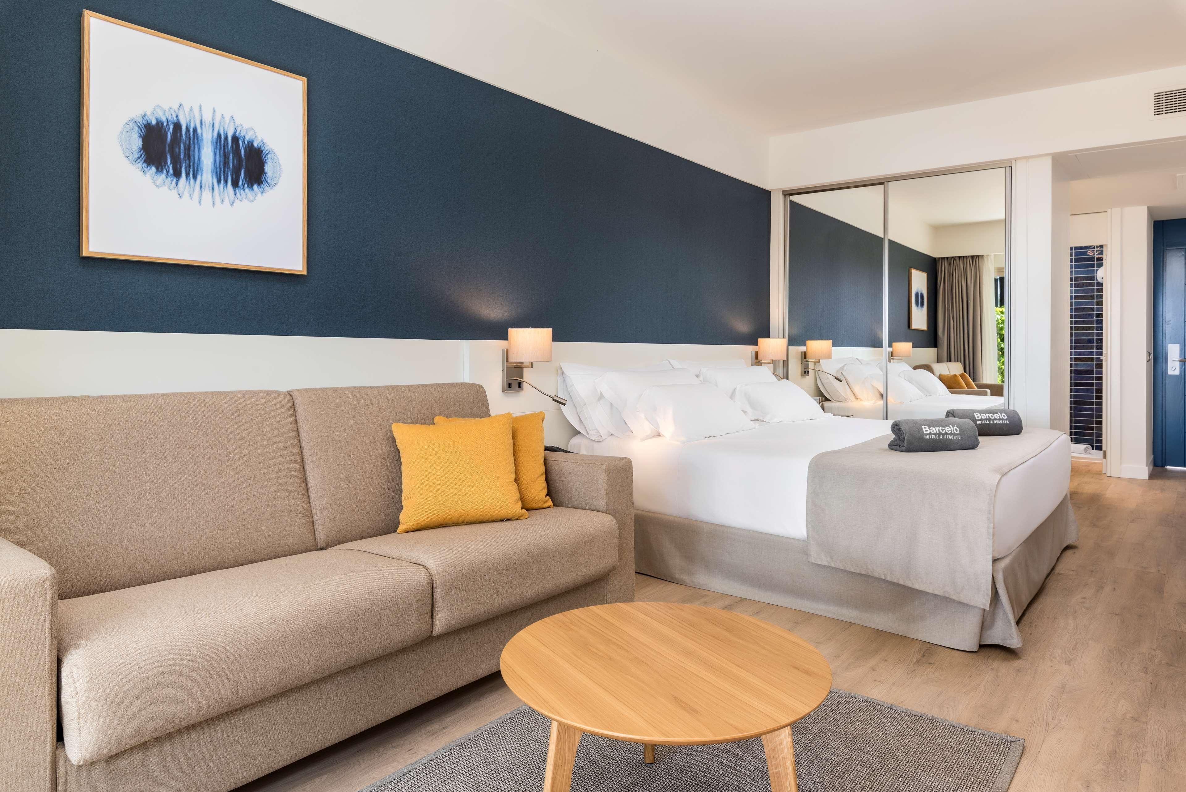 Una habitación de hotel moderna con una cama grande, un sofá, una mesa redonda de madera y una obra de arte enmarcada en la pared.