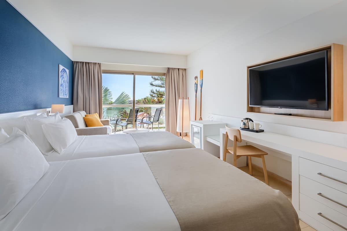 A hotel room with two beds, a desk, a TV, and a balcony with outdoor seating.