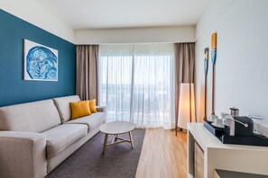 A modern hotel room with a sofa, a small round table, a coffee machine, and a view of the city through sheer curtains.