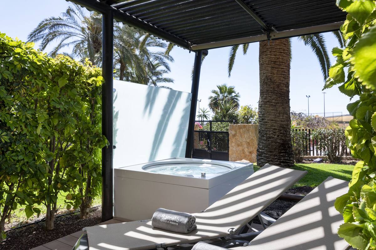 A hot tub under a covered patio with lounge chairs, surrounded by greenery and palm trees.