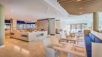 A modern restaurant with a central buffet, seating areas, and a high ceiling with unique lighting.