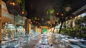 Un restaurante con terraza, mesas dispuestas con manteles blancos y platos verdes, y macetas colgantes con plantas.