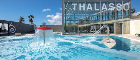 Un área de piscina moderna con un edificio de cristal al fondo, que dice "THALASSO".