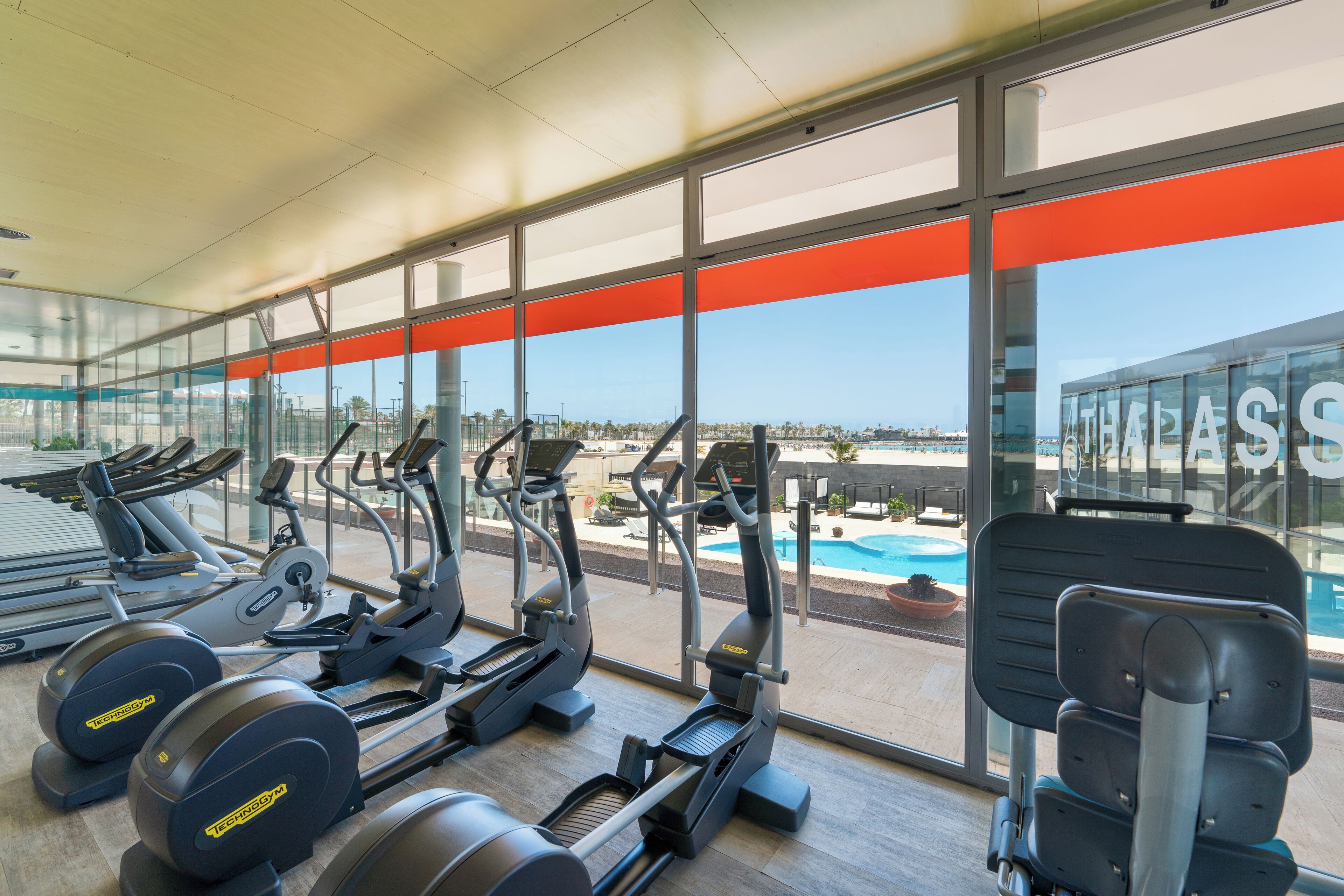A gym with treadmills, elliptical machines, and weight machines, overlooking a pool and a clear sky.
