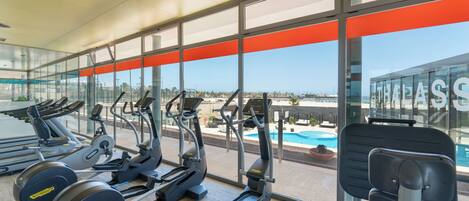A gym with treadmills, elliptical machines, and weight machines, overlooking a pool and a clear sky.