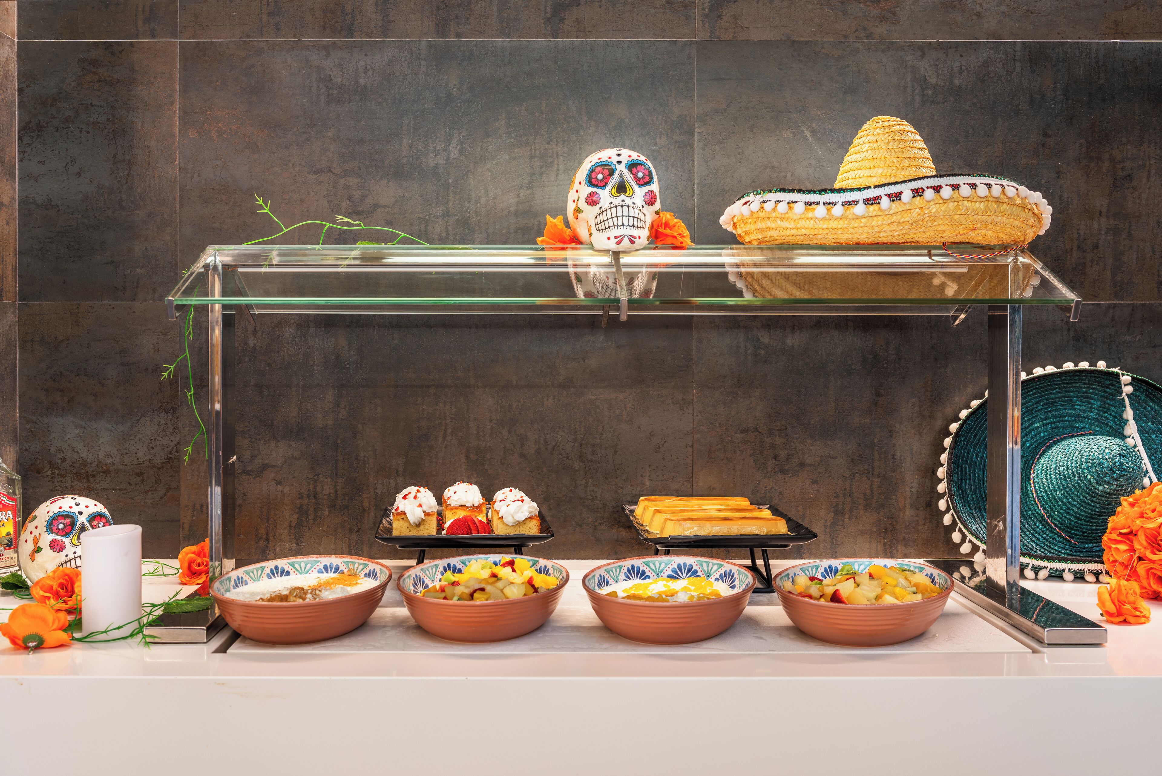 A glass display case with a sombrero, sugar skull, and various desserts.