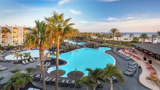 Zona de piscina en un resort con tumbonas, palmeras y vistas al océano.