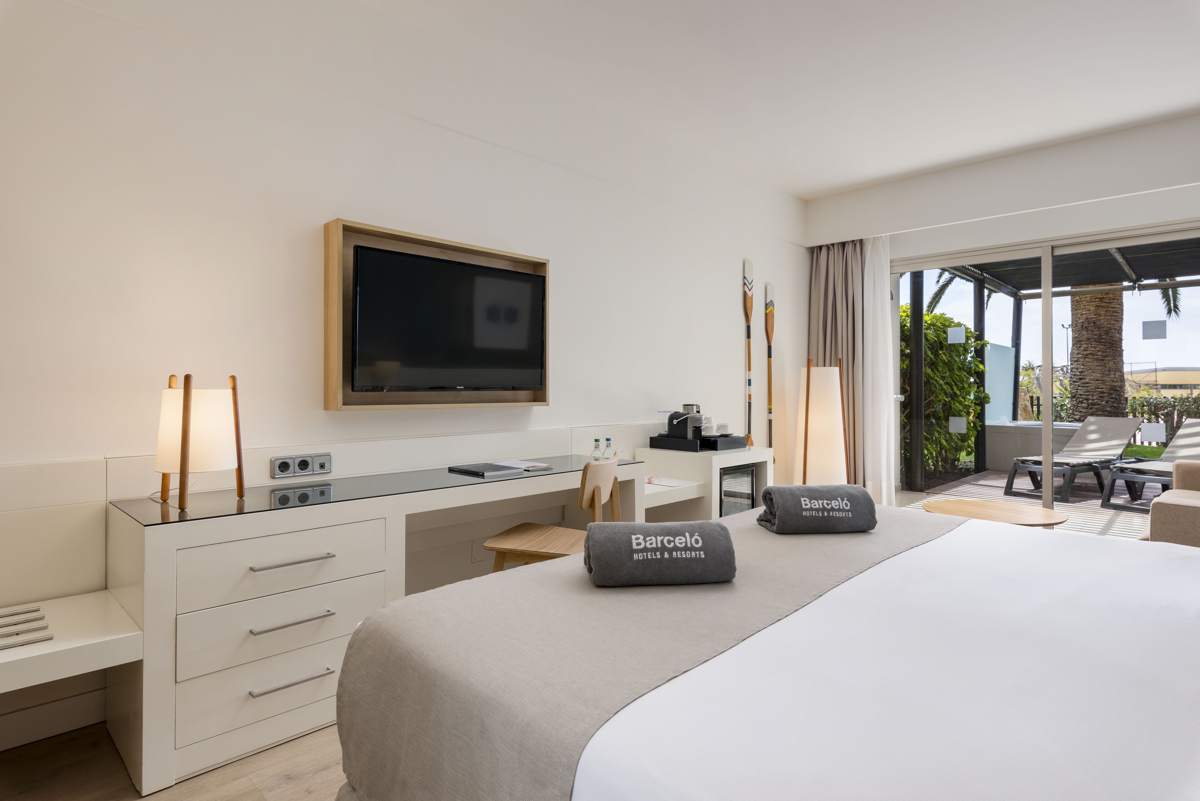 A hotel room with a bed, a desk, a TV, and a balcony with seating.