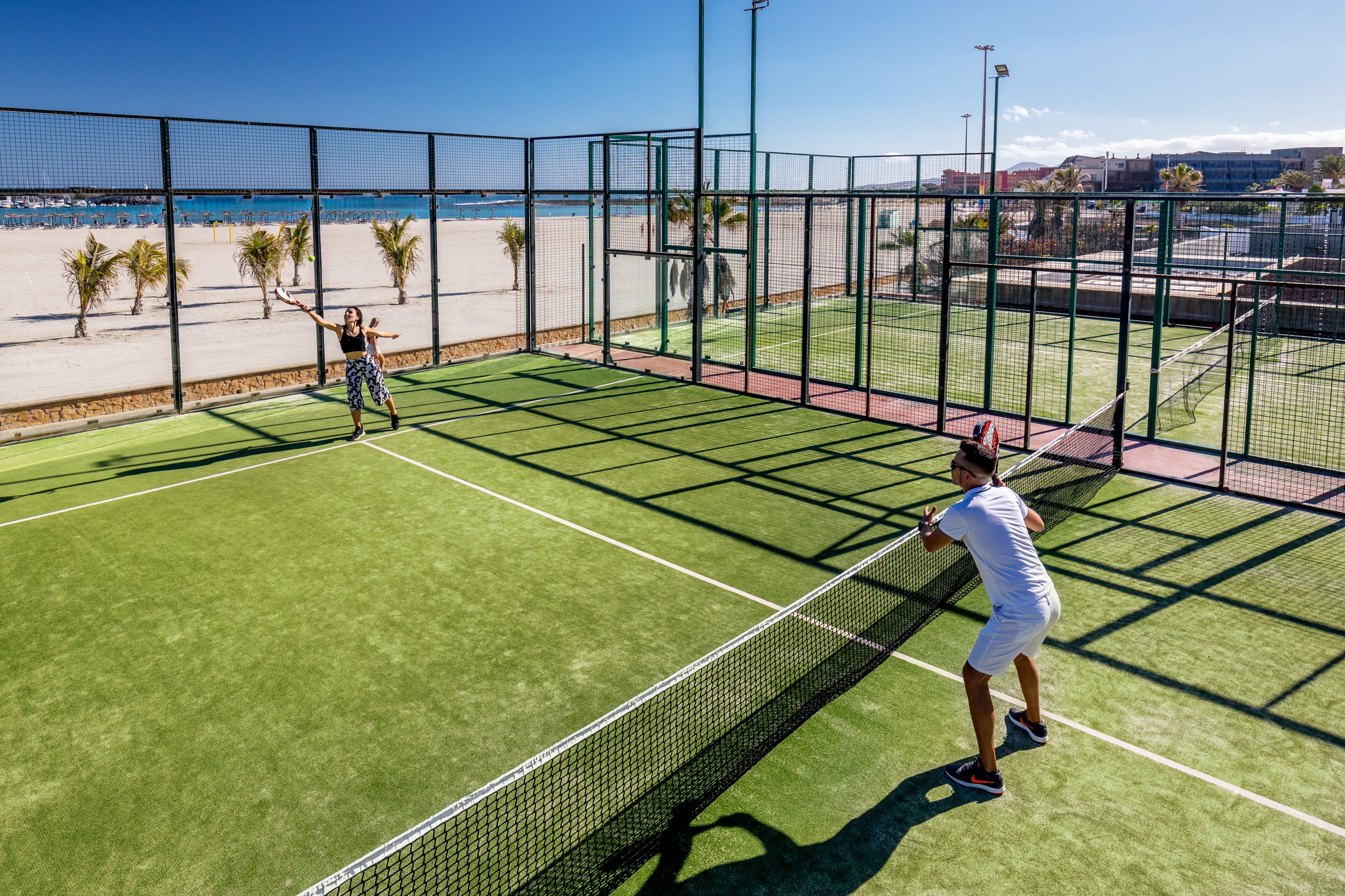 Una cancha de tenis con red, rodeada por una valla, y una playa al fondo.