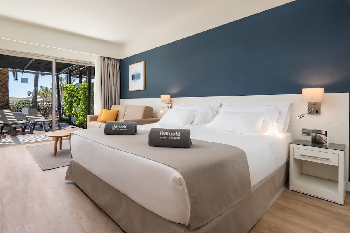 A hotel room with a bed, a nightstand, a chair, a table, and a view of the outdoors.