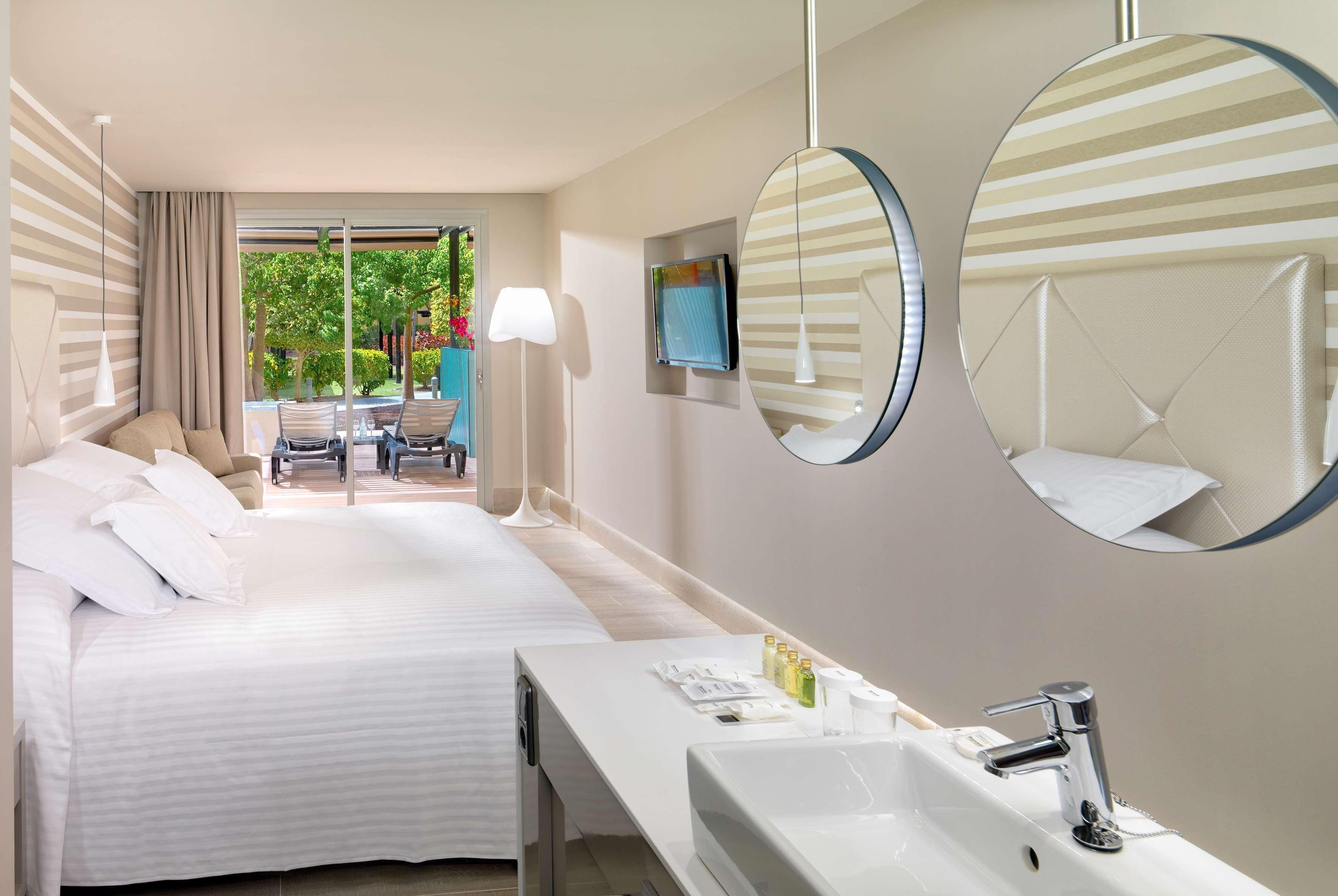 Une chambre d’hôtel avec un lit, une salle de bain comprenant un lavabo et un miroir, et une vue sur un jardin depuis la fenêtre.