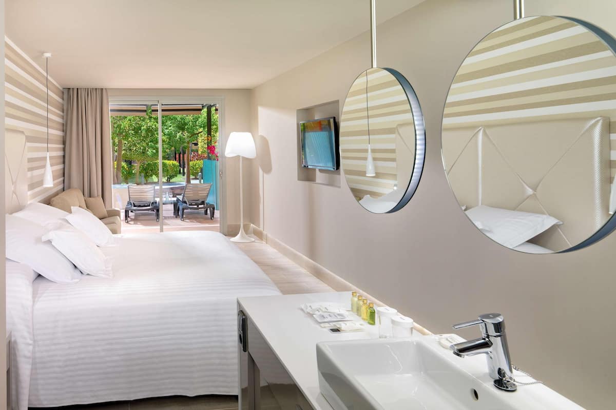 Une chambre d’hôtel avec un lit, une salle de bain comprenant un lavabo et un miroir, et une vue sur un jardin depuis la fenêtre.