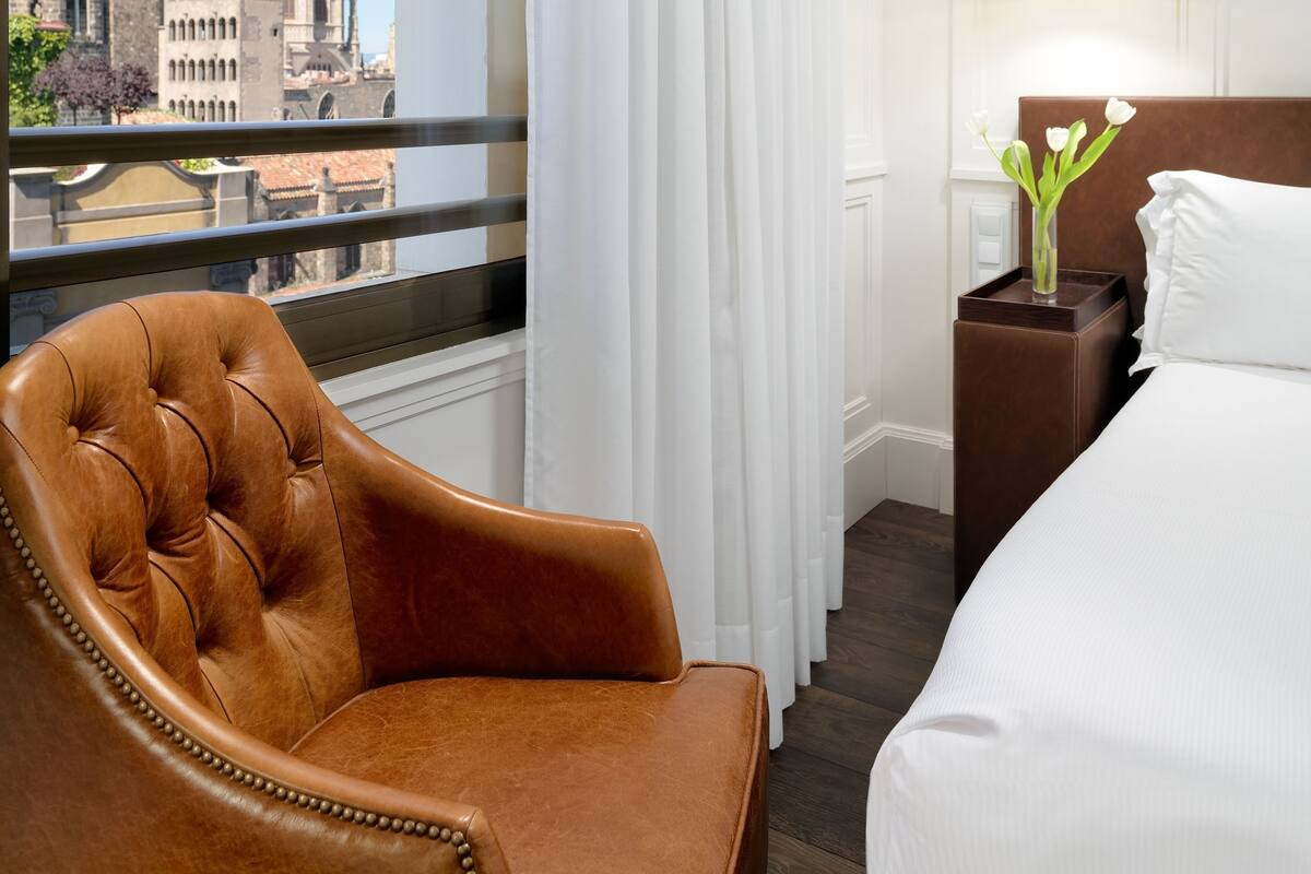A hotel room with a view of a cathedral, a brown leather chair, a bedside table with a vase of flowers, and a hanging light fixture.