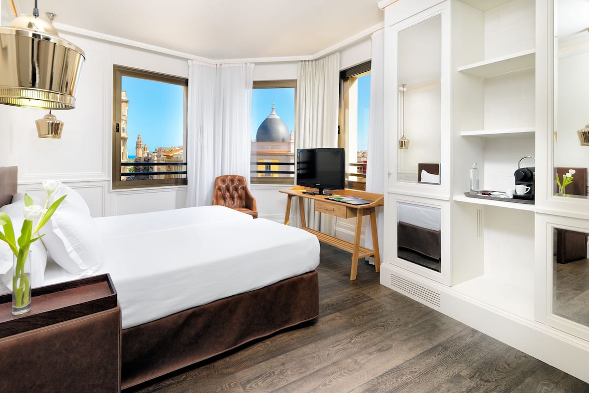 A hotel room with a bed, a desk with a computer, a TV, and a view of a building through the window.