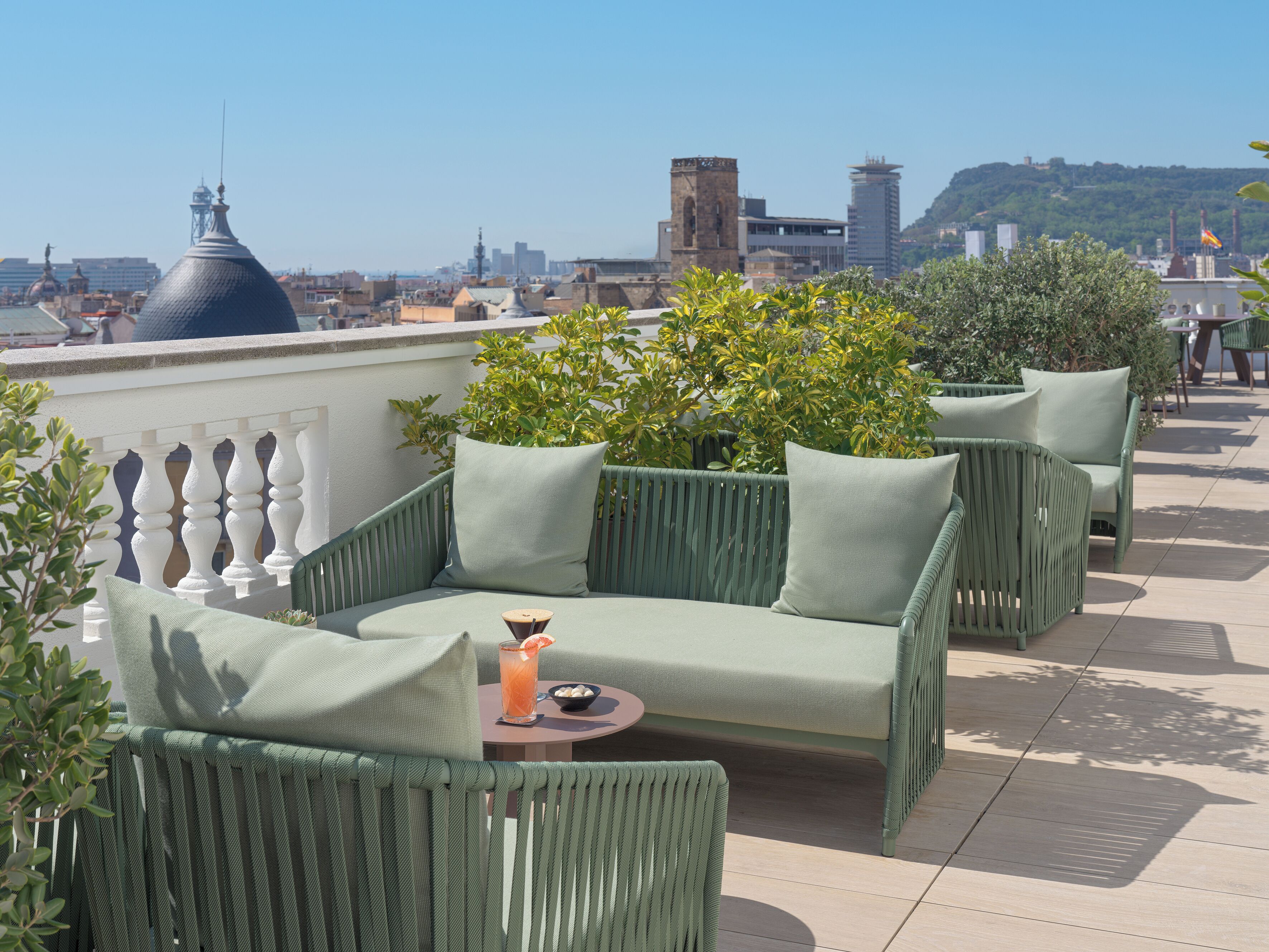 A rooftop terrace with green wicker furniture, a table with a drink, and a view of the cityscape.