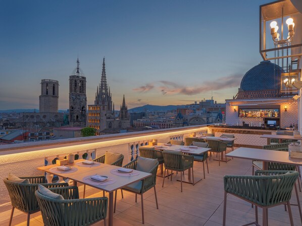 A rooftop bar with tables and chairs, a bar counter with bottles, and a city skyline in the background.