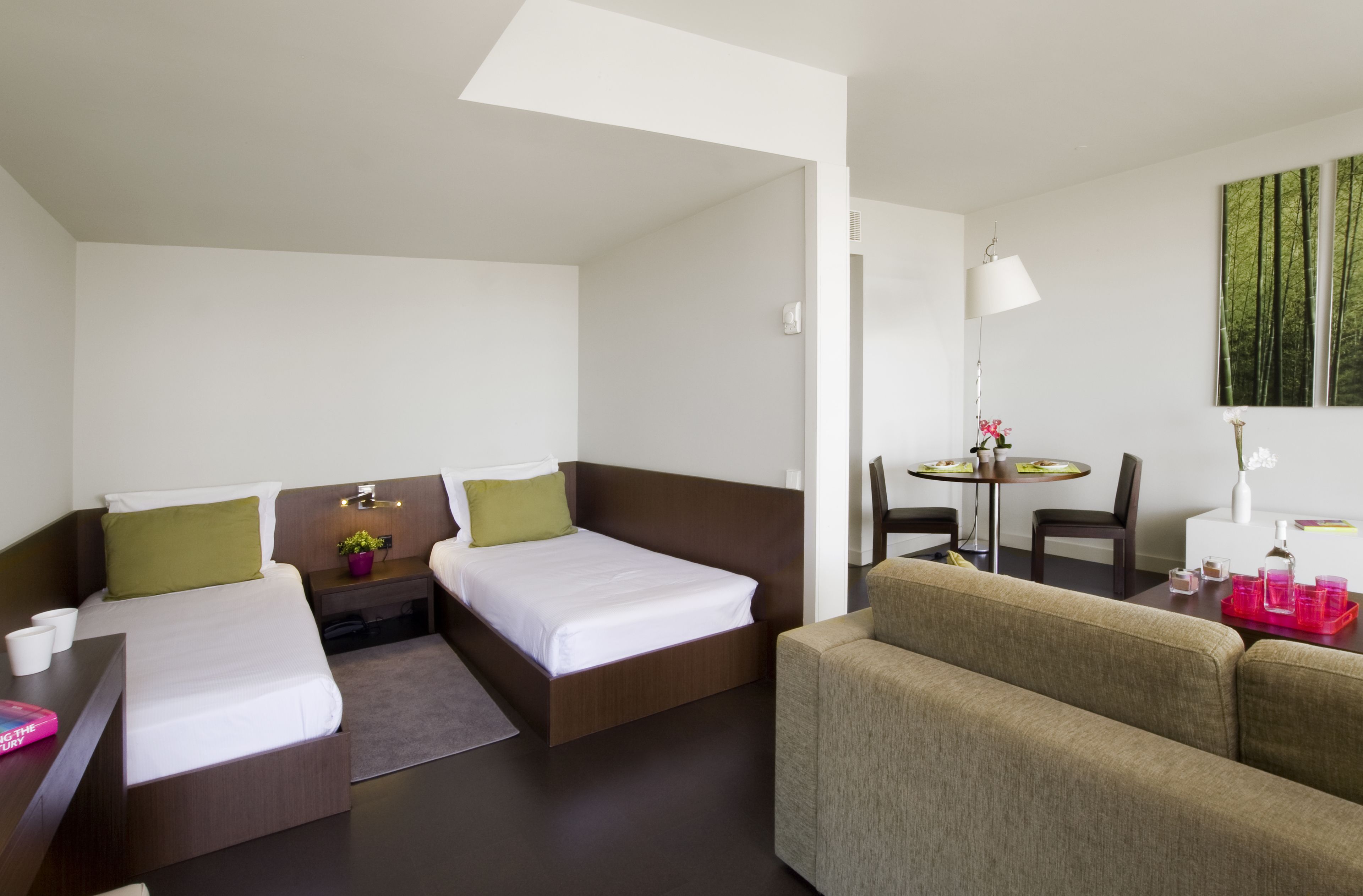 A hotel room with two single beds, a sofa, a small dining table with chairs, and a painting on the wall.