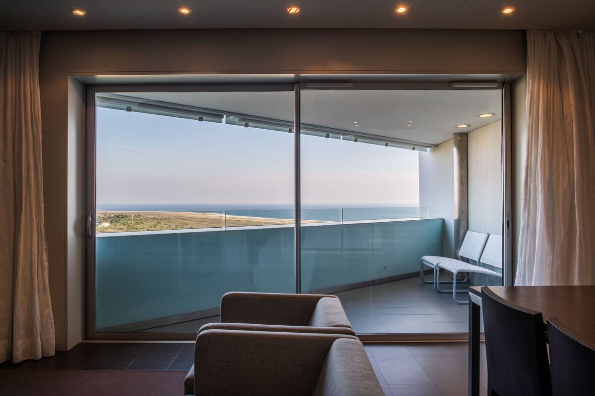 Habitación de hotel moderna con un amplio ventanal corredero que da a un balcón con vista al mar. Cuenta con dos zonas de estar con cojines y una pequeña mesa.