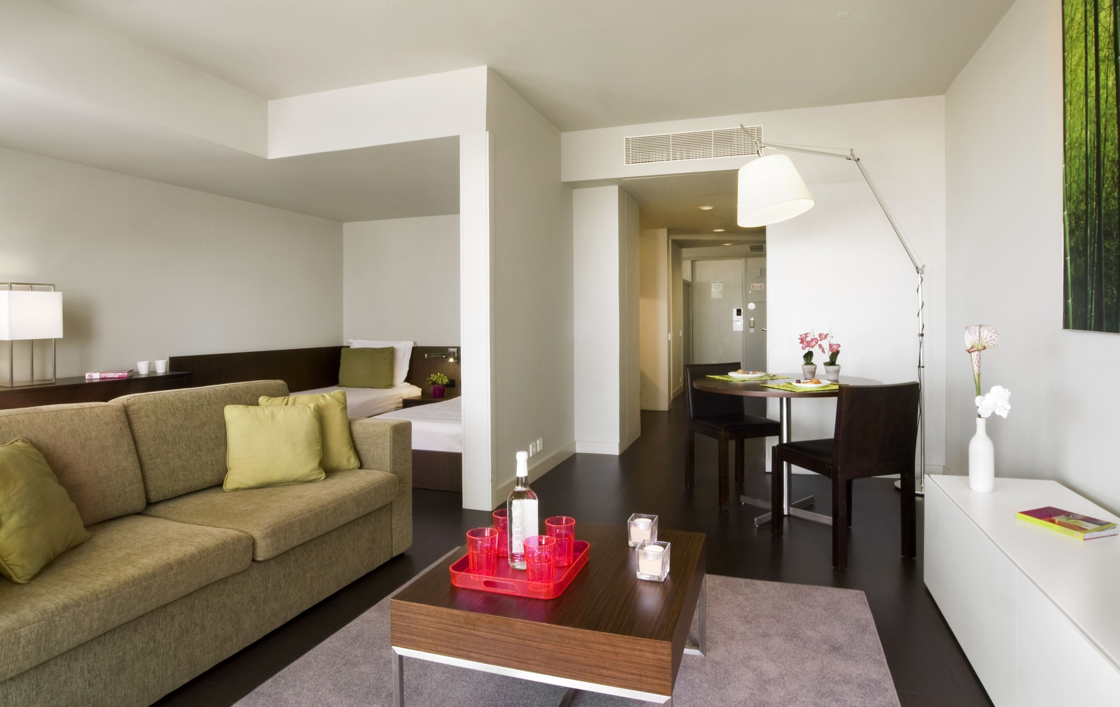 A modern hotel room with a sofa, a dining table with chairs, a coffee table, and a lamp.