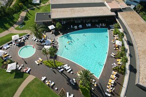 An aerial view of a hotel with a large swimming pool, sun loungers, and a covered area with umbrellas.
