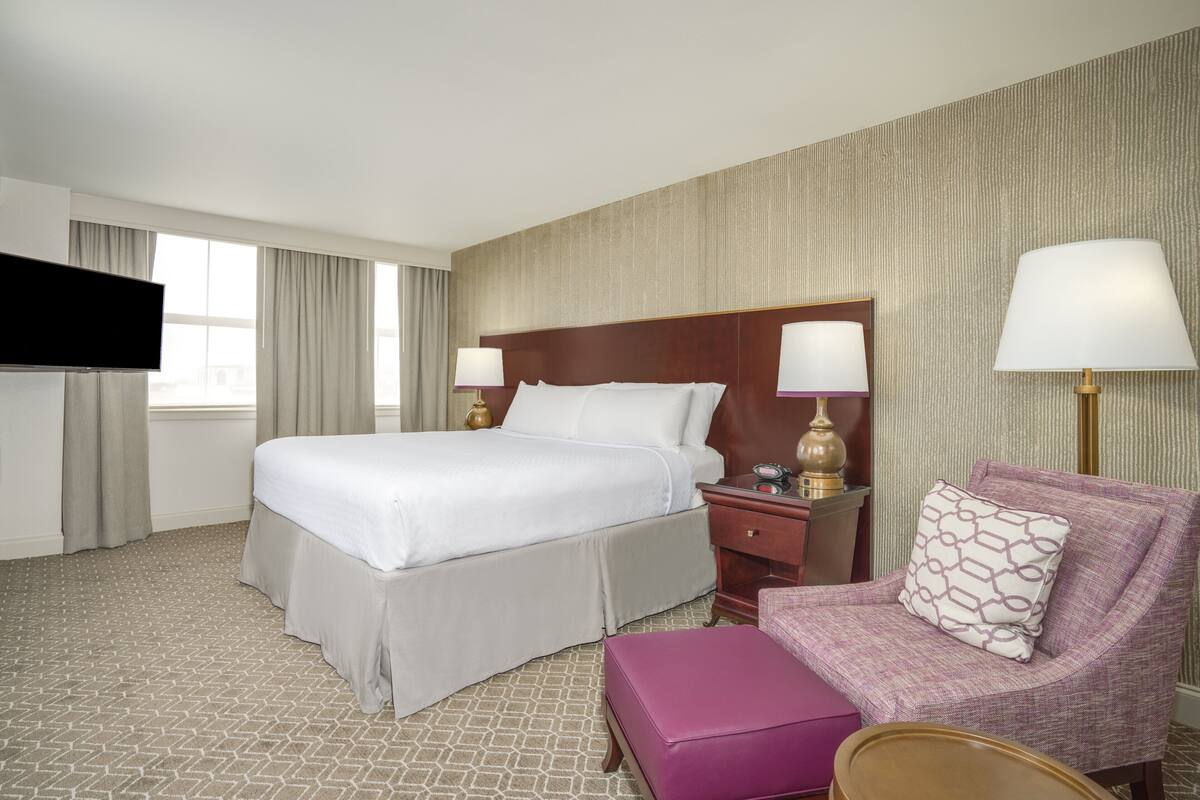 Quarto de hotel com uma cama grande, mesa de cabeceira com luminárias, uma cadeira e televisão.