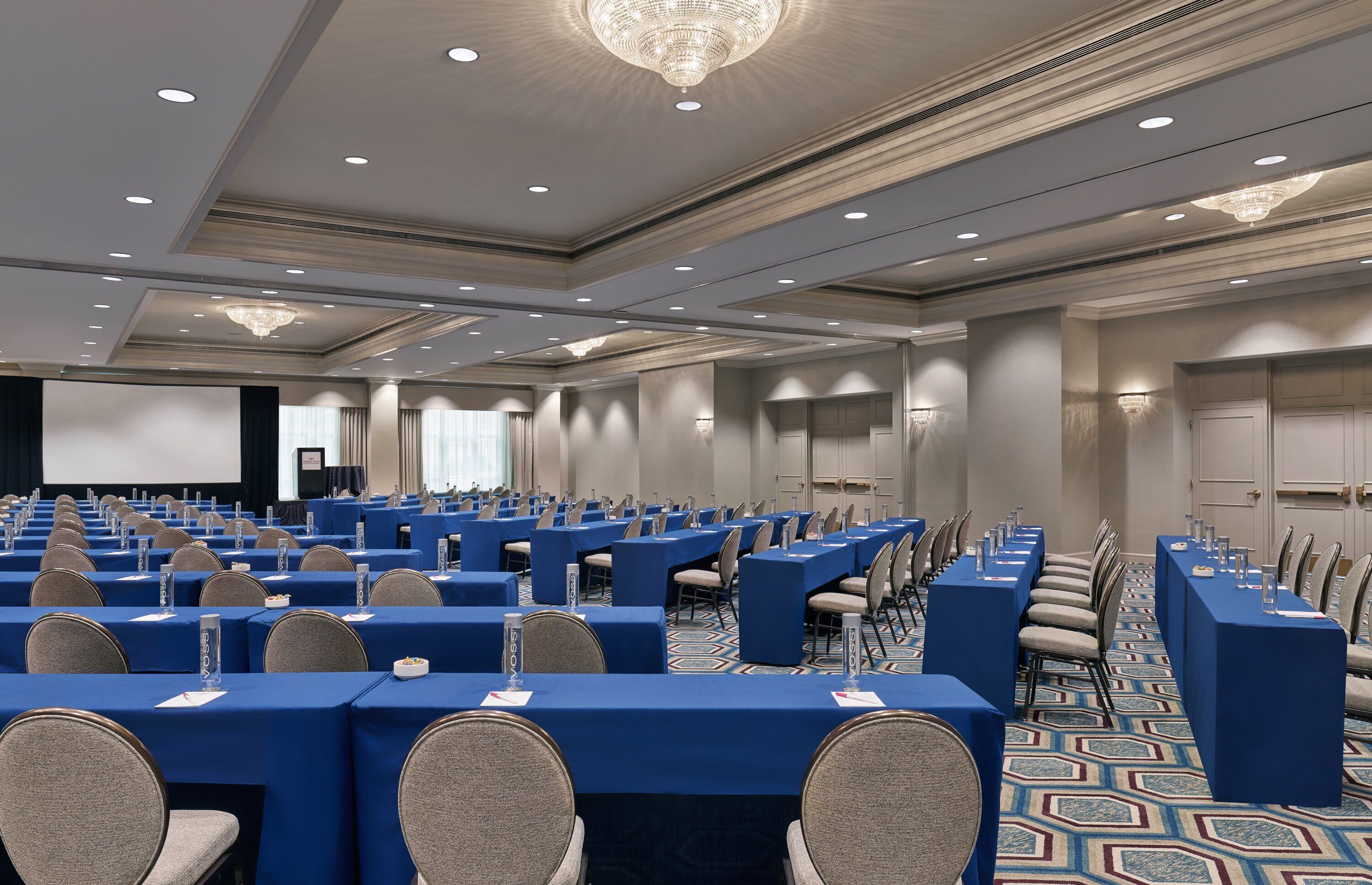 A conference room with rows of tables and chairs, a large screen, and a chandelier.