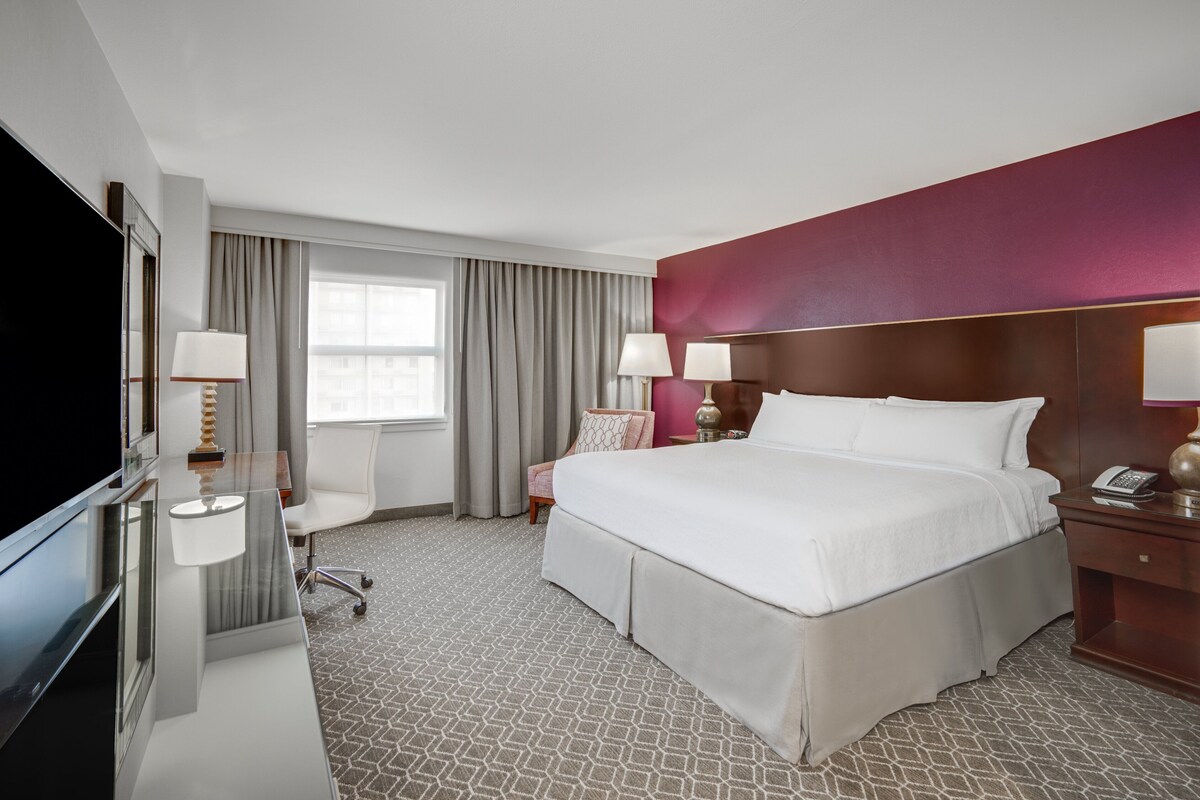 Een hotelkamer met een groot bed, een televisie, een bureau met lamp, een stoel en een raam met gordijnen.