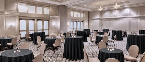 Une grande salle de conférence dotée de nombreuses tables rondes, chacune recouverte d’une nappe noire et agrémentée de bougies blanches. La salle est ornée d’un tapis à motifs, de lustres et de grandes fenêtres à vitres dépolies.